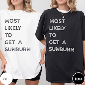 Puede incluir: Una camiseta blanca y una camiseta negra, cada una con el texto MOST LIKELY TO GET A SUNBURN impreso en una fuente oscura y desgastada. Ambas camisetas tienen cuello redondo y mangas cortas, presentadas juntas para comparar.