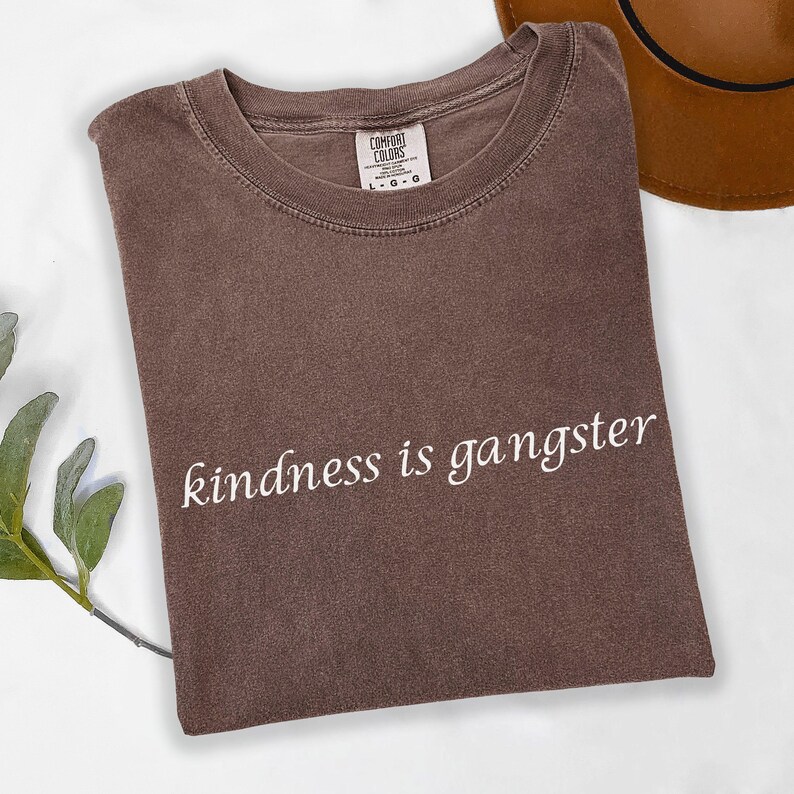May include: A brown Comfort Colors t-shirt with the phrase "kindness is gangster" in white script. The shirt is folded and has a crew neck. A brown hat is partially visible in the upper right corner.