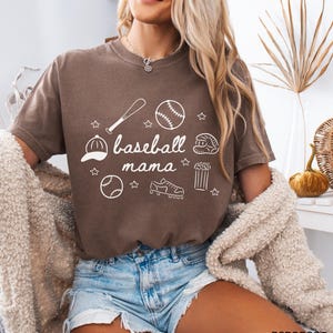 Puede incluir: Una camiseta marr&oacute;n con un dise&ntilde;o gr&aacute;fico blanco que dice "baseball mama" e incluye ilustraciones con tem&aacute;tica de b&eacute;isbol como una pelota de b&eacute;isbol, un bate, un guante y tacos.