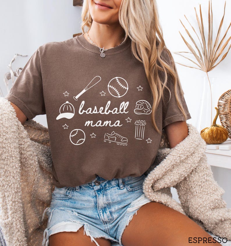 Puede incluir: Una camiseta marr&oacute;n con un dise&ntilde;o gr&aacute;fico blanco que dice "baseball mama" e incluye ilustraciones con tem&aacute;tica de b&eacute;isbol como una pelota de b&eacute;isbol, un bate, un guante y tacos.