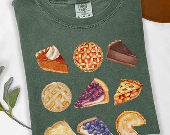 May include: A moss green short-sleeve t-shirt featuring a graphic print of nine various pie slices and whole pies, including pumpkin, lattice-crust fruit, and chocolate varieties. The inner neck label reads Comfort Colors with size L-G-G.