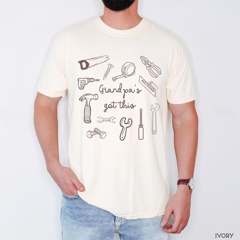 Peut inclure: T-shirt ivoire avec un motif d'outils dessin&eacute;s &agrave; la main, dont une scie, un marteau et une cl&eacute;. Le texte "Grandpa's got this" est imprim&eacute; dans un style manuscrit. Le t-shirt est en mati&egrave;re souple.