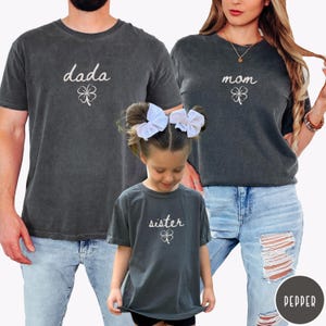 May include: A family wearing matching dark gray t-shirts with white text and a four-leaf clover design. The shirts say "dada", "mom", and "sister". The family is wearing blue jeans.