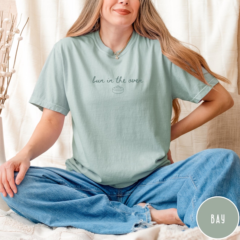 K&ouml;nnte beinhalten: Hellgr&uuml;nes T-Shirt mit dem Schriftzug "bun in the oven" in Schreibschrift. Das Shirt hat eine kleine Abbildung eines Br&ouml;tchens. Das Model tr&auml;gt eine blaue Jeans.