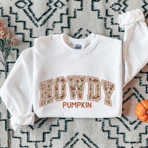 May include: A white long-sleeve crewneck sweatshirt featuring the word HOWDY in large block letters with a tan and brown western-themed pattern inside. Below the word HOWDY, the word PUMPKIN is printed in a solid orange font on the chest area.