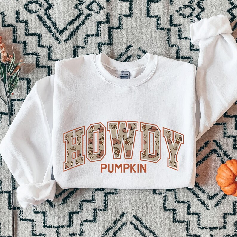 May include: A white long-sleeve crewneck sweatshirt featuring the word HOWDY in large block letters with a tan and brown western-themed pattern inside. Below the word HOWDY, the word PUMPKIN is printed in a solid orange font on the chest area.