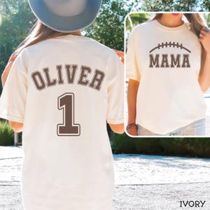 May include: An ivory t-shirt featuring a brown graphic on the back with the name OLIVER and the number 1 in a collegiate font. The front of the shirt displays a brown graphic of a football above the text MAMA in a matching collegiate font.