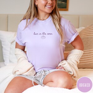K&ouml;nnte beinhalten: Ein helllila T-Shirt mit dem Aufdruck "bun in the oven" in dunkelvioletter Schrift. Das Shirt hat hochgekrempelte &Auml;rmel und ist aus einem weichen Material. Das Shirt ist Teil einer Umstandsmode-Kollektion.