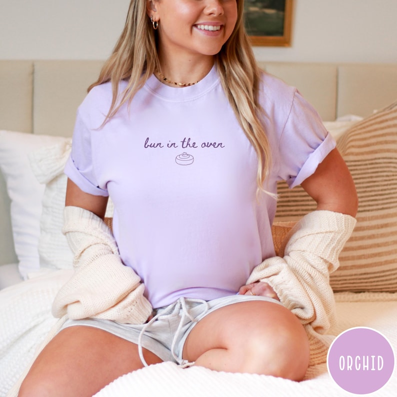 K&ouml;nnte beinhalten: Ein helllila T-Shirt mit dem Aufdruck "bun in the oven" in dunkelvioletter Schrift. Das Shirt hat hochgekrempelte &Auml;rmel und ist aus einem weichen Material. Das Shirt ist Teil einer Umstandsmode-Kollektion.