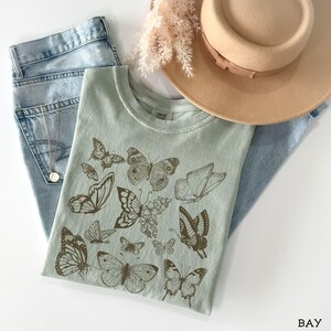 May include: A light green t-shirt with a brown butterfly design. The shirt has a crew neck and short sleeves. The butterflies are arranged in a random pattern across the front of the shirt.