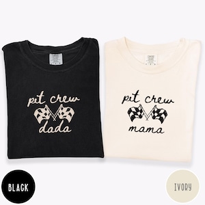 May include: Two t-shirts, one black and one ivory, with the words "pit crew" and crossed checkered flags printed above "dada" and "mama". The shirts are folded and have a crew neck.