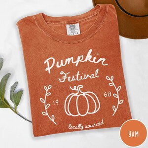 May include: A burnt orange t-shirt with a white graphic print that reads Pumpkin Festival, 1968, and locally sourced. The design features a line-art pumpkin in the center flanked by two leafy vine illustrations. The shirt has a Comfort Colors label.