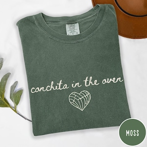 May include: A moss green t-shirt with the phrase "conchita in the oven" in white script, accompanied by a white heart-shaped graphic. The shirt is folded and has a Comfort Colors tag.