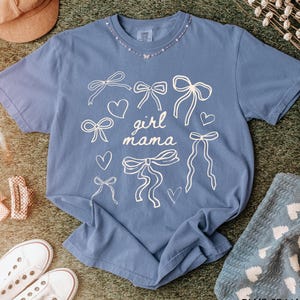 May include: A blue t-shirt with a white graphic design of bows and hearts. The text "girl mama" is printed on the shirt.