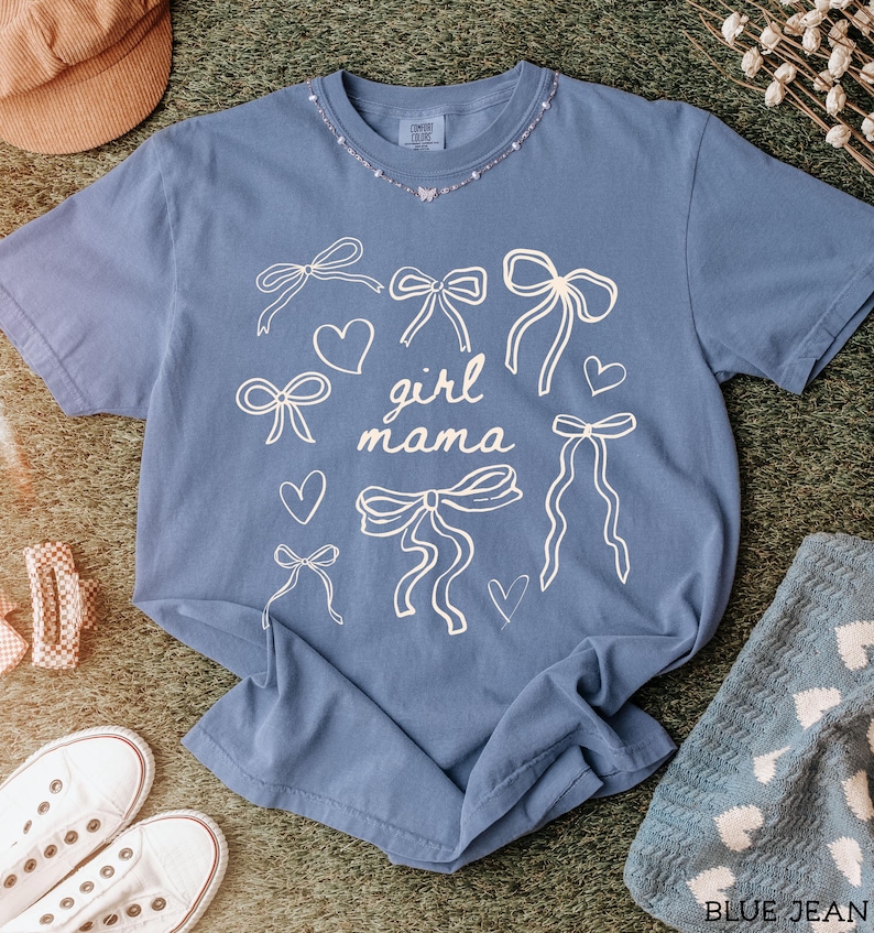 May include: A blue t-shirt with a white graphic design of bows and hearts. The text "girl mama" is printed on the shirt.