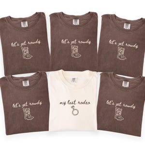May include: Five brown t-shirts and one white t-shirt. The brown shirts feature the text "let's get rowdy" above a line-art cowboy boot graphic. The white shirt features the text "my last rodeo" above a line-art diamond ring graphic.