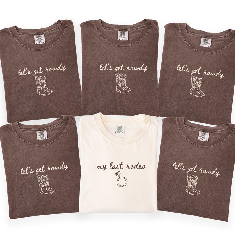May include: Five brown t-shirts and one white t-shirt. The brown shirts feature the text "let's get rowdy" above a line-art cowboy boot graphic. The white shirt features the text "my last rodeo" above a line-art diamond ring graphic.