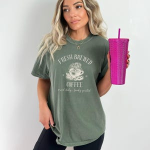 Op de afbeelding: Een olijfgroen T-shirt met korte mouwen en een witte afbeelding van een dampende koffiekop met de tekst "FRESH BREWED COFFEE" en "served daily - family operated". Een felroze beker met textuur en een rietje wordt in de hand gehouden.