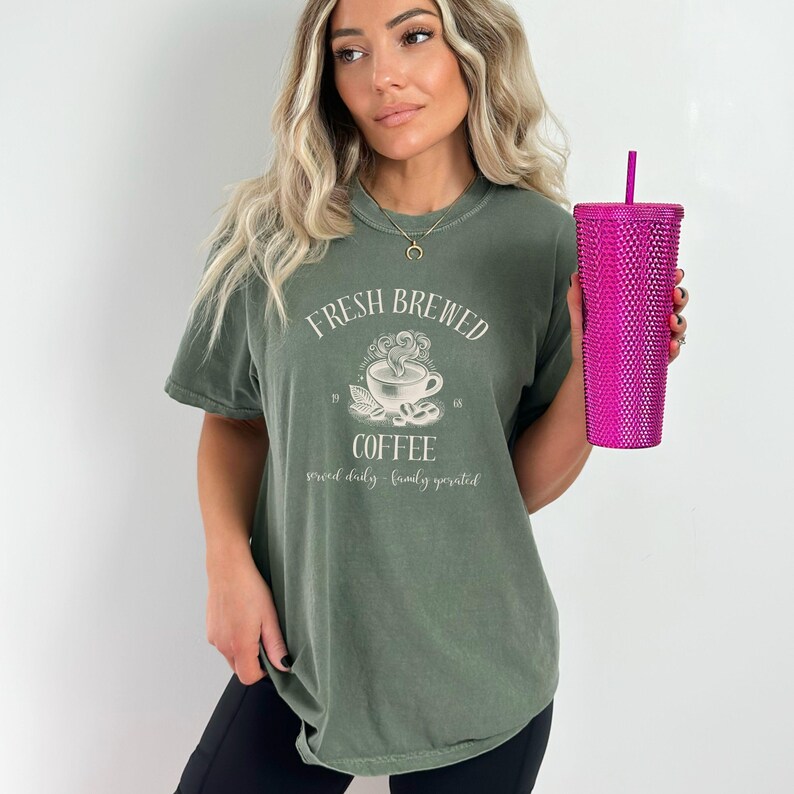 Op de afbeelding: Een olijfgroen T-shirt met korte mouwen en een witte afbeelding van een dampende koffiekop met de tekst "FRESH BREWED COFFEE" en "served daily - family operated". Een felroze beker met textuur en een rietje wordt in de hand gehouden.