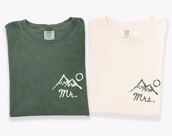 May include: Two crew neck t-shirts, one in moss green and one in ivory. The moss shirt features a white graphic of mountains, a circle, and the text 'Mr.' The ivory shirt features a moss green graphic of mountains, a circle, and the text 'Mrs.'