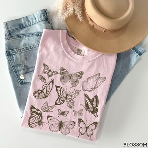May include: A pink t-shirt with a brown butterfly design. The shirt has the text "Comfy Cozy Club" printed on the front.