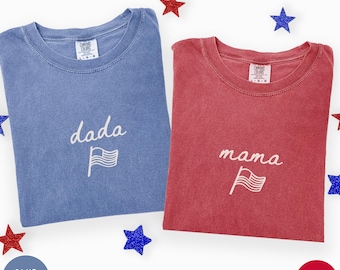 May include: Two crew neck t-shirts, one in blue jean and one in crimson, each featuring white embroidered text and a flag graphic. The blue shirt displays the word "dada" and the red shirt displays the word "mama" with a small flag icon below.