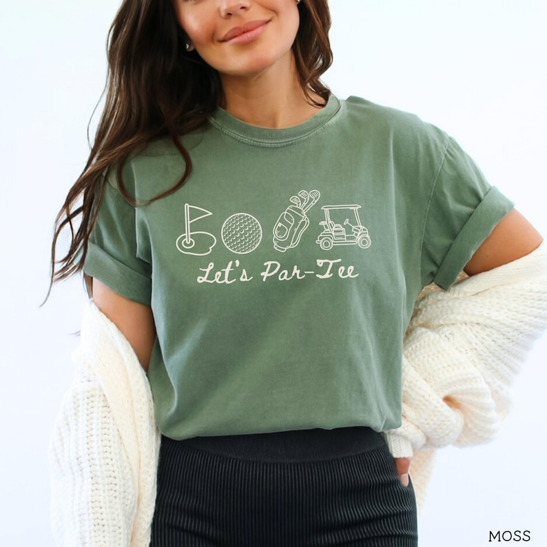 May include: Moss green t-shirt with white graphic of golf-related icons: flag, golf ball, golf bag, and golf cart. The text "Let's Par-Tee" is below the icons. The shirt is made of a soft material.