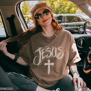 May include: Espresso-colored t-shirt with the word "JESUS" in a stylized font and a cross graphic. The shirt is worn by a person wearing sunglasses and a brown hat. The image is taken inside a car.