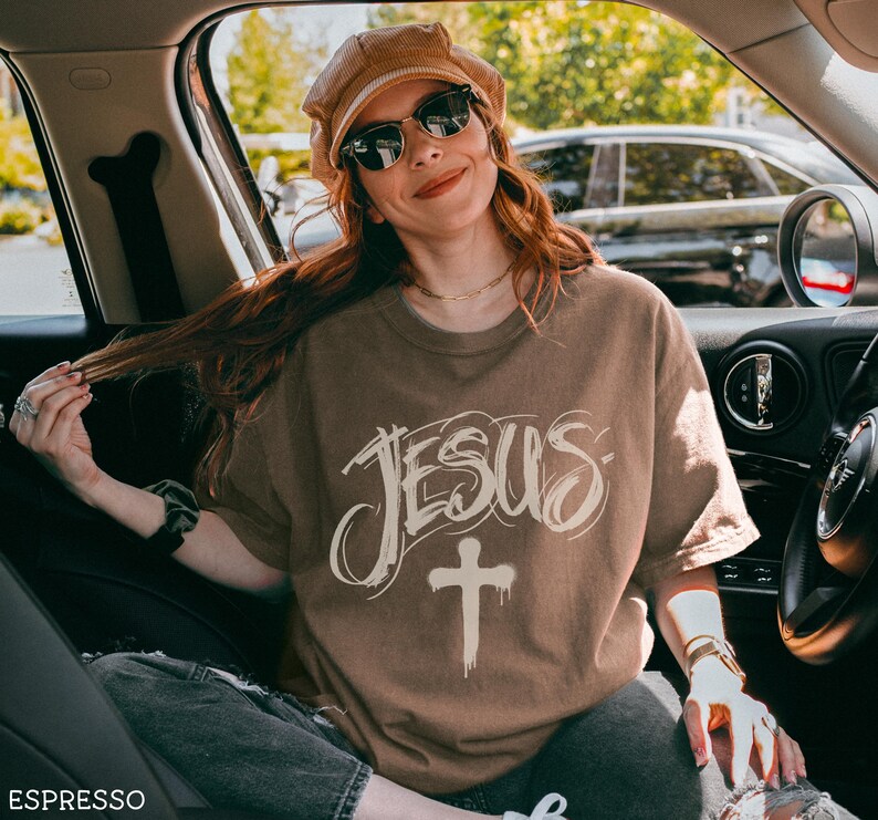 May include: Espresso-colored t-shirt with the word "JESUS" in a stylized font and a cross graphic. The shirt is worn by a person wearing sunglasses and a brown hat. The image is taken inside a car.