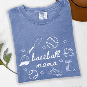 Puede incluir: Una camiseta azul claro con un dise&ntilde;o blanco de tem&aacute;tica de b&eacute;isbol. El dise&ntilde;o incluye un bate de b&eacute;isbol, una pelota de b&eacute;isbol, un guante de b&eacute;isbol, una gorra de b&eacute;isbol y el texto "baseball mama".