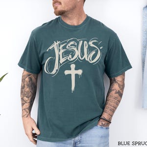 May include: Blue spruce t-shirt with the word "JESUS" in a stylized font and a cross graphic. The shirt is a solid color and has a crew neck. The text "BLUE SPRUCE" is at the bottom right.