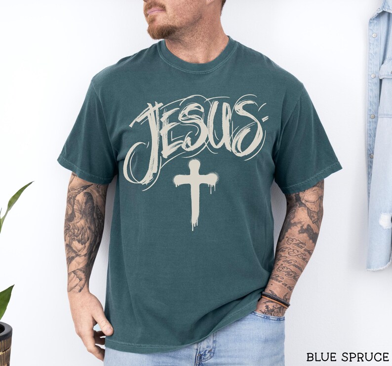 May include: Blue spruce t-shirt with the word "JESUS" in a stylized font and a cross graphic. The shirt is a solid color and has a crew neck. The text "BLUE SPRUCE" is at the bottom right.