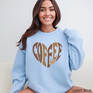 May include: Light blue sweatshirt with a heart-shaped design that says "COFFEE" in brown and tan lettering. A small tan heart is below the word. The sweatshirt is a solid light blue color.