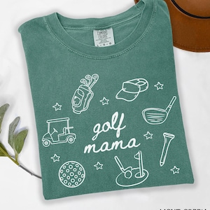 Puede incluir: Camiseta verde claro con un dise&ntilde;o gr&aacute;fico blanco que dice "golf mama" e incluye im&aacute;genes relacionadas con el golf como palos de golf, un carrito de golf, una pelota de golf y un sombrero de golf.