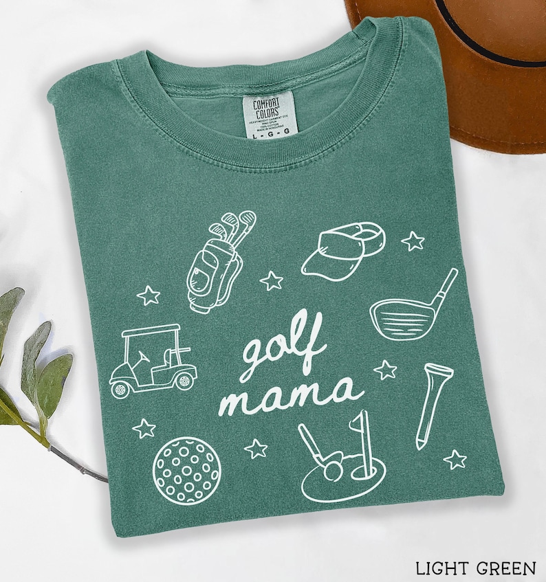Puede incluir: Camiseta verde claro con un dise&ntilde;o gr&aacute;fico blanco que dice "golf mama" e incluye im&aacute;genes relacionadas con el golf como palos de golf, un carrito de golf, una pelota de golf y un sombrero de golf.