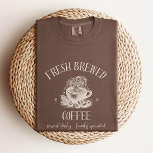 Op de afbeelding: Een bruin t-shirt met een witte afbeelding van een dampende koffiekop en de tekst FRESH BREWED COFFEE, 1968, en served daily - family operated. Het t-shirt heeft een Comfort Colors merklabel aan de binnenkant van de ronde halslijn.