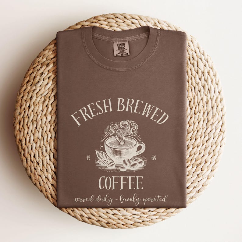 Op de afbeelding: Een bruin t-shirt met een witte afbeelding van een dampende koffiekop en de tekst FRESH BREWED COFFEE, 1968, en served daily - family operated. Het t-shirt heeft een Comfort Colors merklabel aan de binnenkant van de ronde halslijn.
