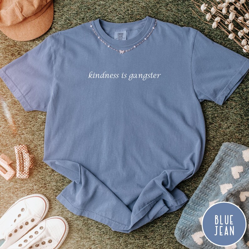May include: A blue jean colored short-sleeve t-shirt with the phrase "kindness is gangster" printed on the front. The shirt has a crew neck and is accessorized with a delicate necklace. The shirt is displayed on a green surface with other accessories.