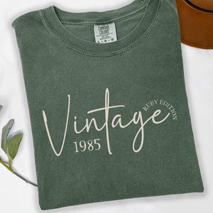 May include: A moss green t-shirt with the word "Vintage" in a cursive font, the year "1985", and the words "Ruby Edition". The shirt is folded and has a tag that says "Comfort Colors". A brown hat is visible in the background.
