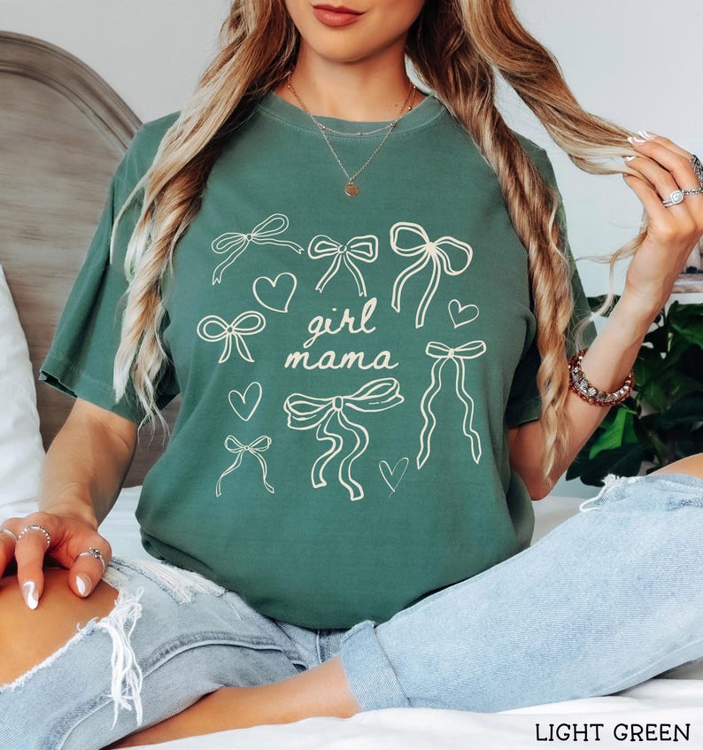 May include: A light green t-shirt with a graphic design of bows and hearts. The text "girl mama" is printed on the shirt.