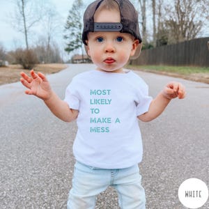Puede incluir: Una camiseta blanca de manga corta con el texto MOST LIKELY TO MAKE A MESS en una fuente de color verde azulado desgastado. La camiseta se combina con pantalones vaqueros azul claro y una gorra de camionero de malla negra y marr&oacute;n.