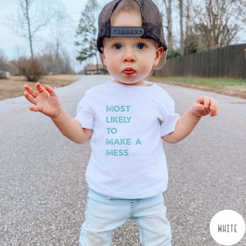 Puede incluir: Una camiseta blanca de manga corta con el texto MOST LIKELY TO MAKE A MESS en una fuente de color verde azulado desgastado. La camiseta se combina con pantalones vaqueros azul claro y una gorra de camionero de malla negra y marr&oacute;n.