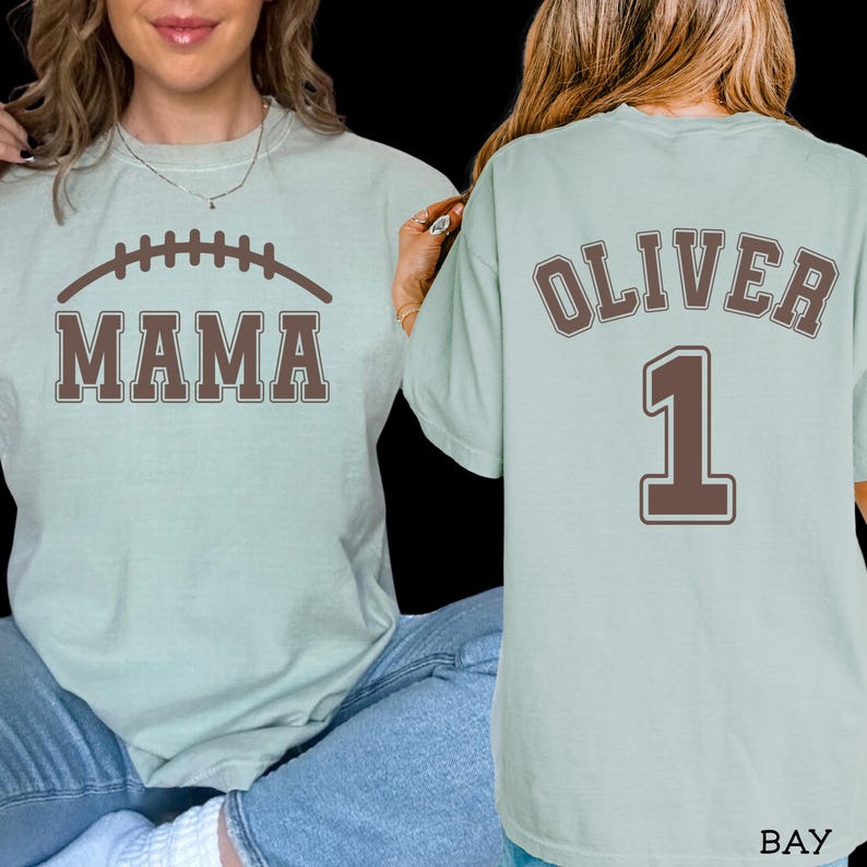 May include: A light green t-shirt featuring a brown graphic on the front with a football lace design above the word MAMA. The back of the t-shirt displays the name OLIVER in a brown collegiate font above the large brown number 1.
