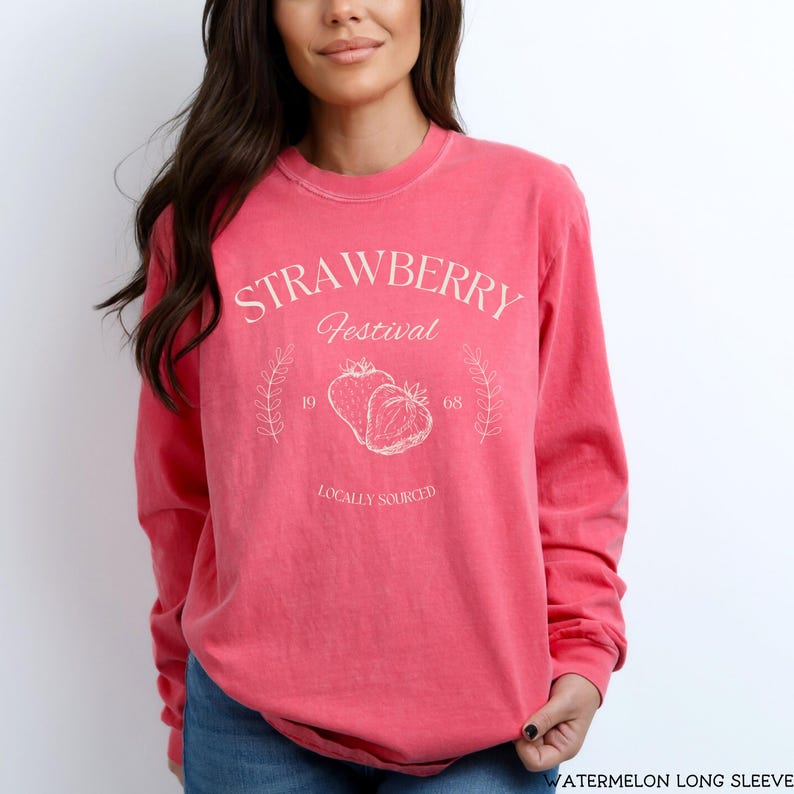 K&ouml;nnte beinhalten: Ein pinkfarbenes Langarmshirt mit einem wei&szlig;en Grafikdesign, auf dem "Strawberry Festival" mit zwei Erdbeeren und dem Text "Locally Sourced" steht.