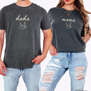 May include: Two matching t-shirts in a dark charcoal color with the words "dada" and "mama" in a cream color. Each shirt has a small graphic of a stroller with a golf club and ball.