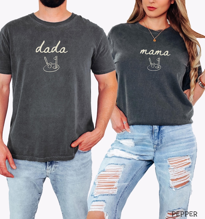 May include: Two matching t-shirts in a dark charcoal color with the words "dada" and "mama" in a cream color. Each shirt has a small graphic of a stroller with a golf club and ball.