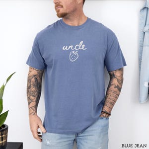 May include: A dusty blue short-sleeved t-shirt featuring the word uncle written in a white cursive script above a simple white line drawing of a strawberry. The shirt has a crew neckline and a relaxed fit, providing a casual look.