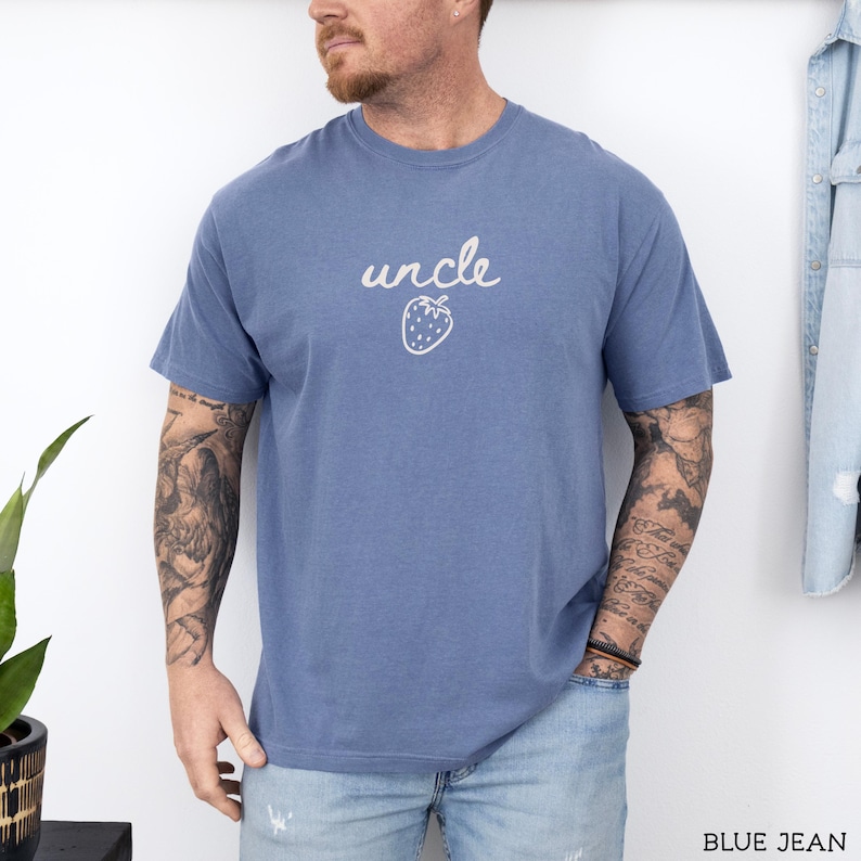 May include: A dusty blue short-sleeved t-shirt featuring the word uncle written in a white cursive script above a simple white line drawing of a strawberry. The shirt has a crew neckline and a relaxed fit, providing a casual look.
