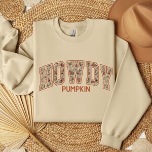 May include: A beige crewneck sweatshirt featuring the text HOWDY PUMPKIN in a collegiate font. The letters of HOWDY are filled with a repeating pattern of brown cowboy hats and horseshoes, while the word PUMPKIN is printed in solid orange.