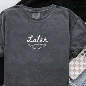 Pu&ograve; includere: Felpa girocollo grigio scuro con la scritta "Later" in corsivo bianco e una grafica di un alligatore bianco. La felpa ha un aspetto vintage e lavato. &Egrave; un capo casual per tutti i giorni. Realizzata in materiale morbido.
